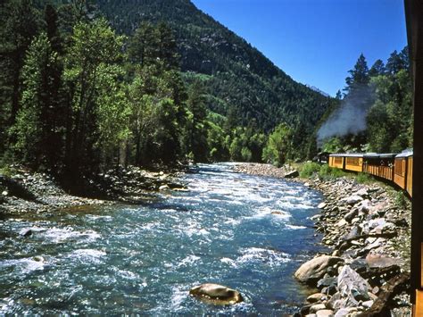 A Train Traveling Down Tracks Next To A River In The Middle Of Trees