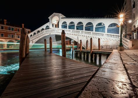 A Wooden Walkway Over Water With Buildings In The Background And Blue